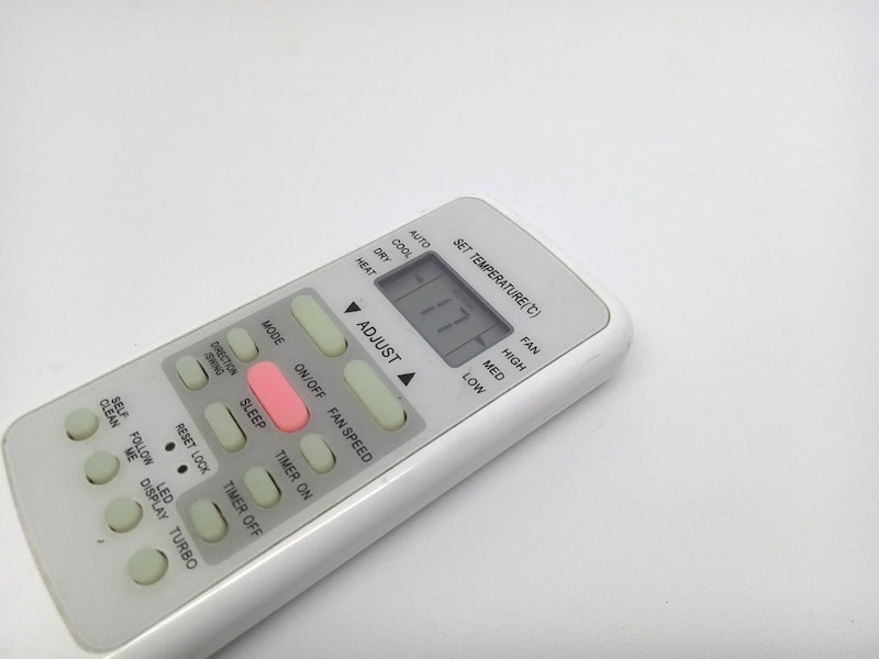 Close-up of a white remote control for air conditioning, featuring various buttons and a small display screen showing the number 77.