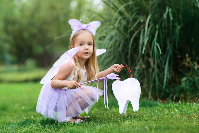 A young child dressed as a fairy, holding a wand and a large model of a tooth, crouches on grass with tall plants in the background.