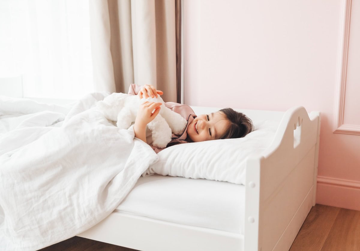 A person lying in bed hugging a plush toy, with white bedding and curtains in a light-colored room.