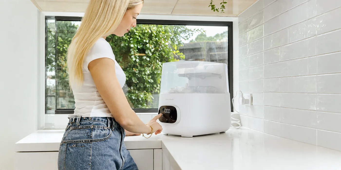 A woman pressing the touch panel on the front of the Pristine Bottle Care Station, which sits on a white kitchen counter loaded with bottles.