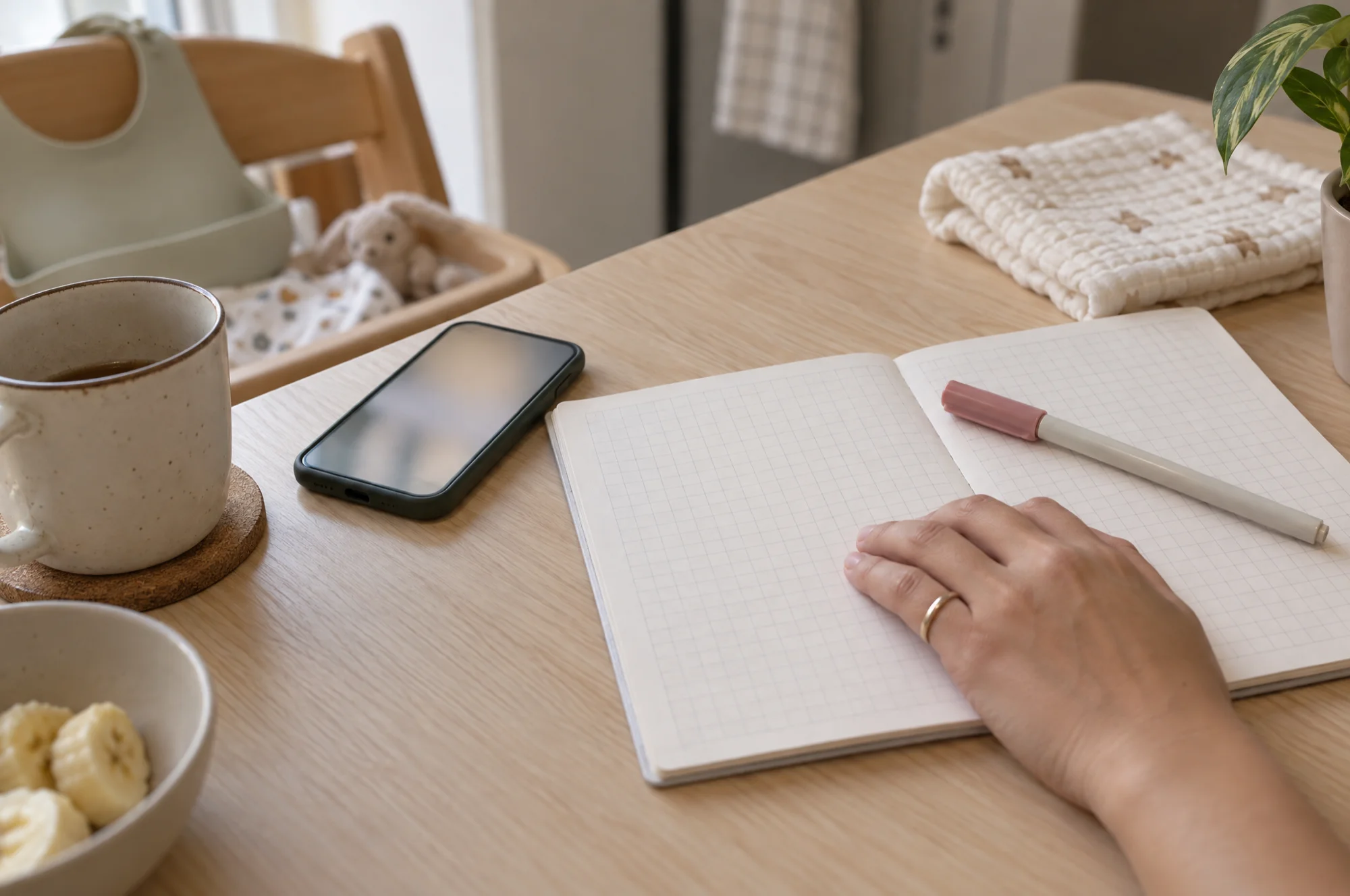 A parent's hand rests on an open booklet at the kitchen table, beside a phone and a pen, ready to log the latest weigh-in.